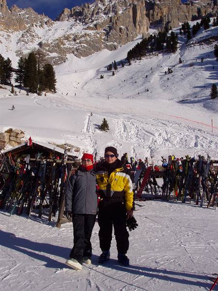 Two skiers by a ski rack in front of snowy Dolomites
