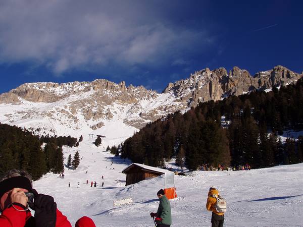 Skiers on snow with wooden huts and snowy Dolomite peaks in the background