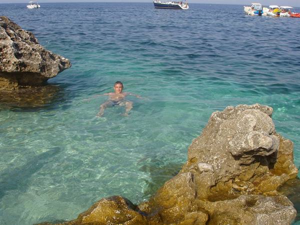 Man swimming between the rocks in turquoise water, boats moored behind