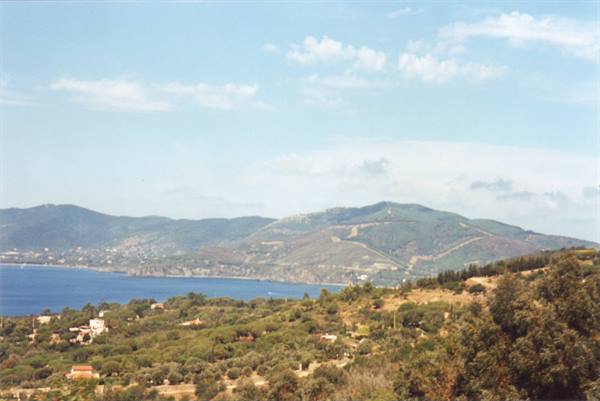 Blue sea between green hills, scattered houses and Mediterranean scrub under cloudy sky