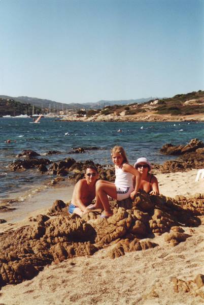 Girl sitting on a rock with her parents, sandy beach and sailboats on blue sea