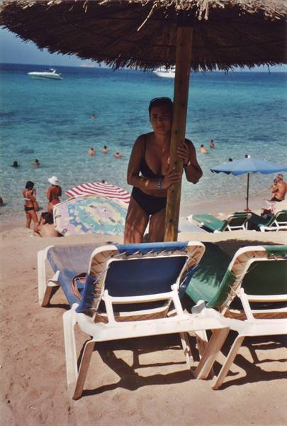 Woman in black swimsuit under straw umbrella, beach with loungers and swimmers