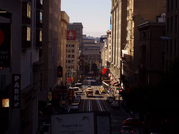 Traffic and pedestrians descend a steep city street between skyscrapers in backlight