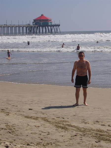 Man in swim trunks on sand, surfers and Huntington Beach pier behind