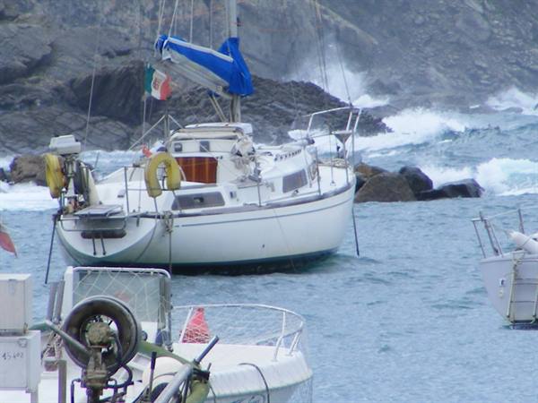 White sailboat anchored among rocks, waves crashing nearby