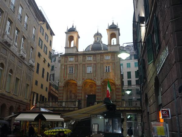 Baroque church with dome amid Genoa buildings, Italian flag and market stalls
