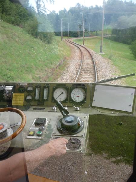 Train driver's cab with gauges and controls, curved tracks through green meadows