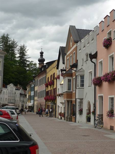Colorful houses with geraniums and a domed tower line a cobbled street under grey sky