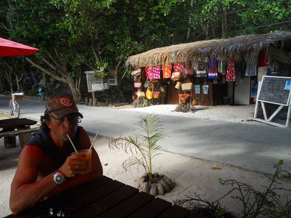 Man in cap sipping a drink outside thatched souvenir stall with hanging bags