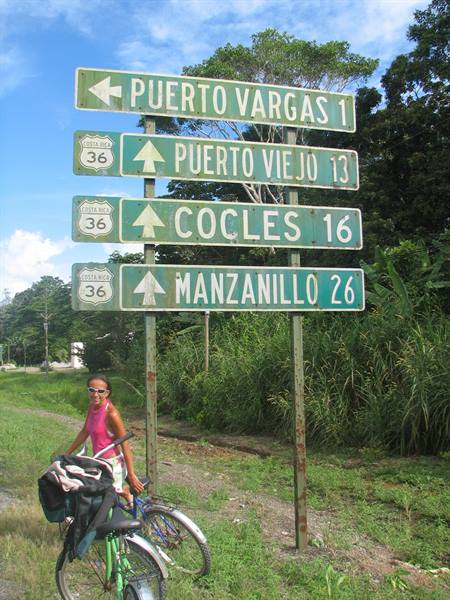 Cyclist in pink top beside green road signs pointing to Puerto Viejo and Manzanillo