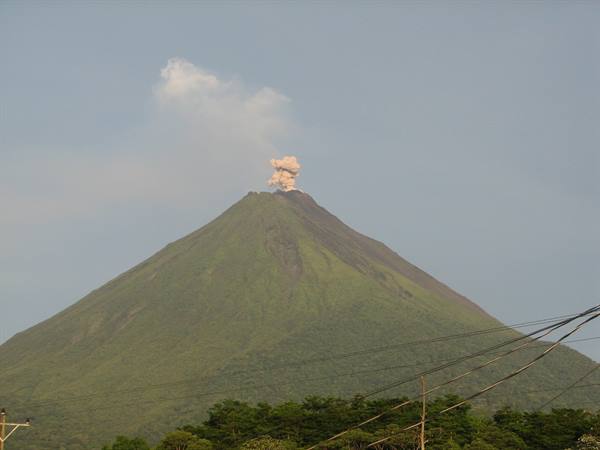 Arenal volcano, green and forested, emits a gray ash puff above power lines
