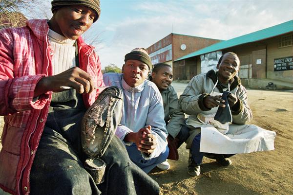 Four men polishing leather shoes on dirt ground in front of warehouses