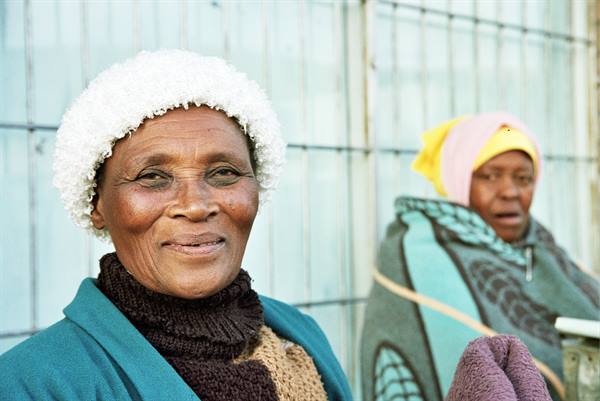 Smiling woman in white hat and shawl in front of a corrugated metal wall