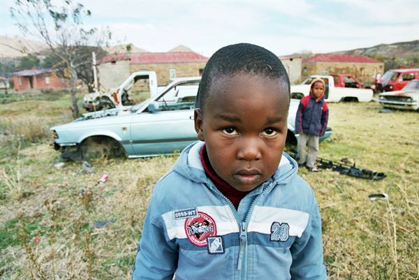 Boy in light blue jacket stares at camera, abandoned cars behind