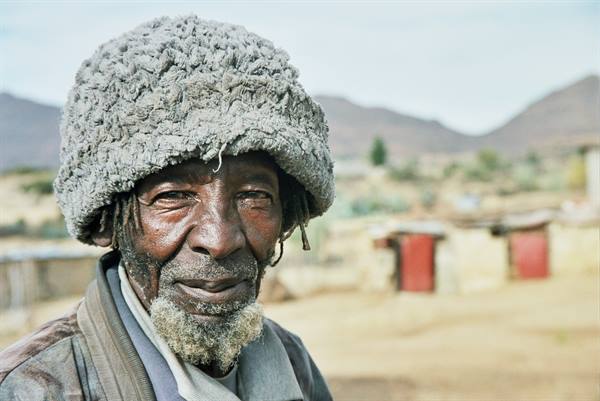 Elderly man in grey woolen hat, village and mountains behind him