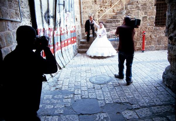 Photographers shoot a bride in white gown and groom on stone steps, cobblestone alley