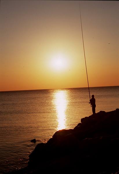 Silhouetted fisherman with rod on rocks, low sun over the sea