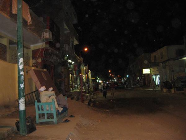 Men sitting on a green wooden bench on a night street, streetlights glowing