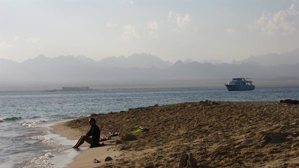 Woman sitting on sand, turquoise sea with boat, hazy mountains behind