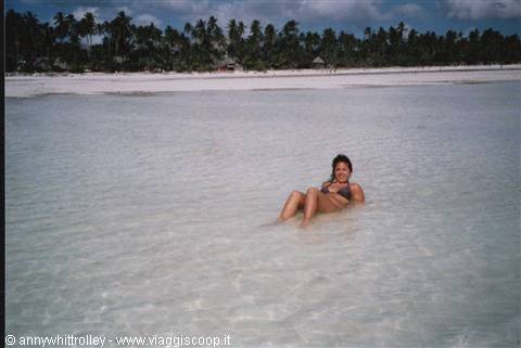 Woman lying in shallow turquoise water, white sandy beach and palm trees behind