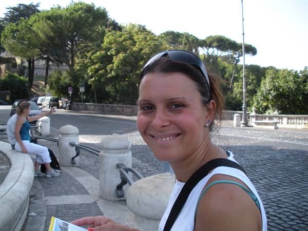Smiling woman with sunglasses on her head near a stone fountain, cobblestones and pines