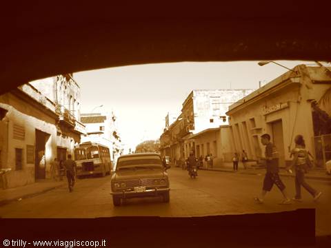 Vintage car driving down a dusty street lined with colonial buildings and pedestrians