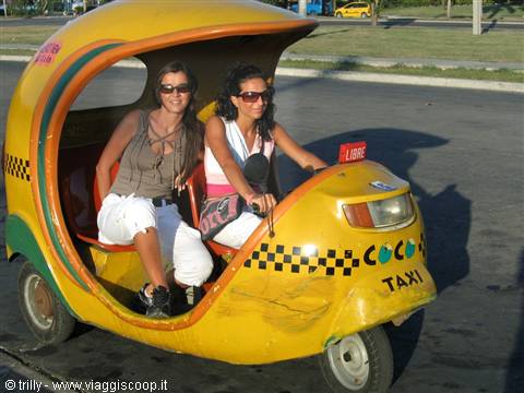 Two women with sunglasses riding a yellow three-wheeled coco taxi on a sunny street