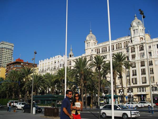 Couple strolling among palms and ornate buildings under blue sky in Alicante