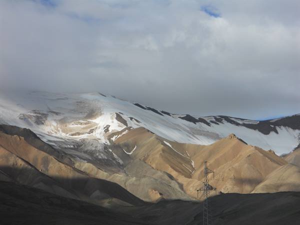 Snowy peak among clouds and brown mountains, electricity pylon in foreground