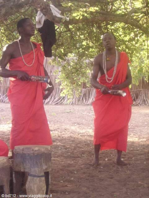 Two men in red robes with shell necklaces blow ivory horns under a tree