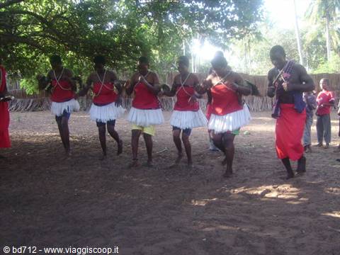 Dancers in white skirts and red tops drumming and stepping on sandy ground