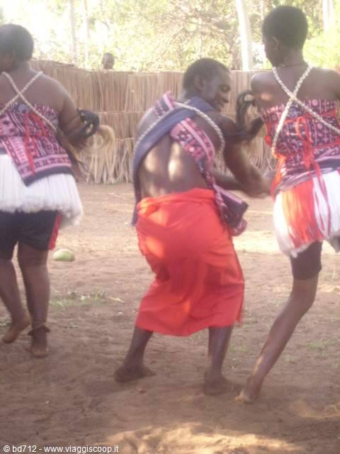 Dancers in red and white traditional skirts dance on bare dirt near huts