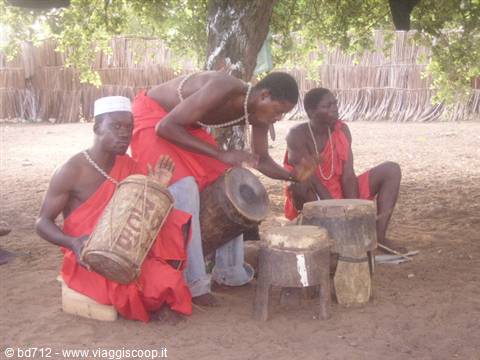 Drummers in red robes play wooden drums under a tree in the village
