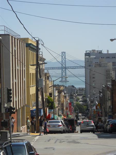 Steep city street with cars and pedestrians, Bay Bridge in the background