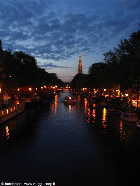 Amsterdam canal at dusk, boat gliding by, Westerkerk tower lit above trees