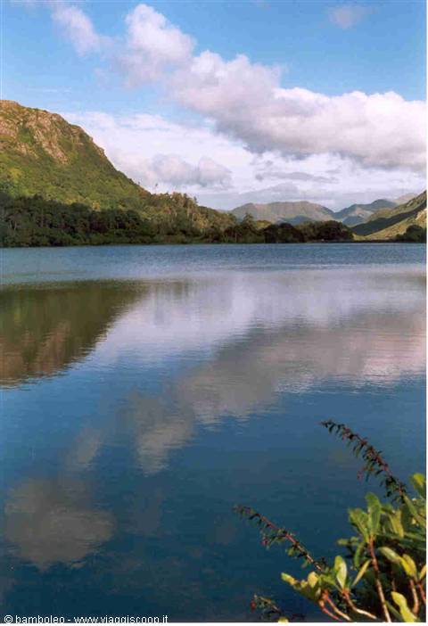 Lake surrounded by green hills and mountains, clouds reflected on calm water