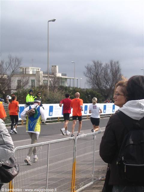 runners in orange shirts past metal barriers, spectators under grey sky