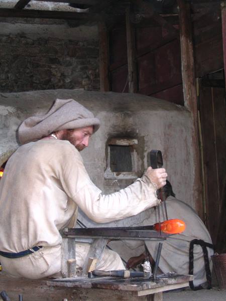 Glassmaker in period costume shapes glowing glass in front of the furnace
