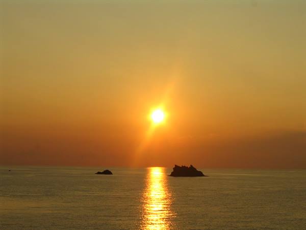 Low sun over calm sea, dark rocks silhouetted, golden reflection on the water