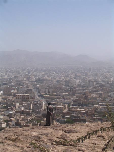 Man in traditional dress on rock overlooking dense city with dry mountains