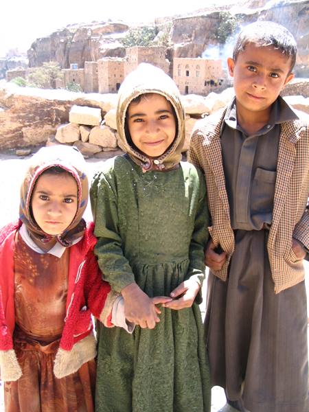 Three children in traditional clothes pose in the sun before old mud-brick houses
