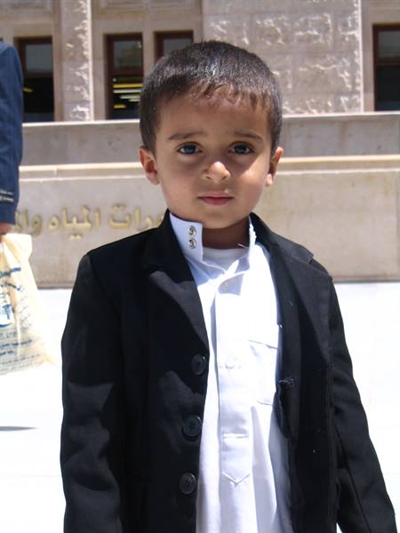 Young boy in black jacket and white shirt before an Arabic-inscribed building