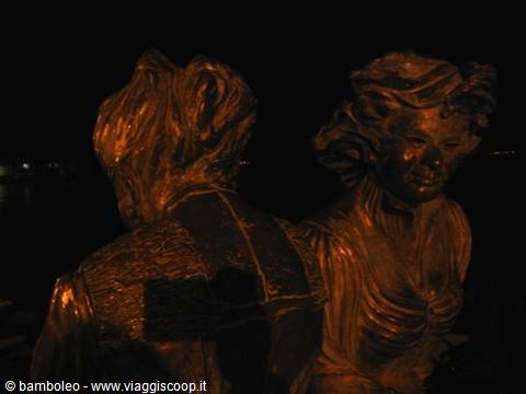 Bronze statue of two female figures lit at night beside the water