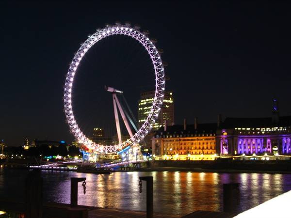 Illuminated ferris wheel at night by the river, purple lights on buildings