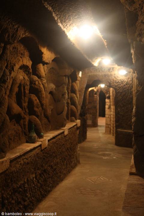 Tuff-carved underground corridor with sculpted walls, warm lamps, bottles