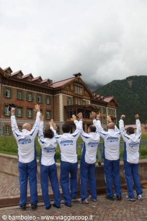 Group in white and blue tracksuits waving in front of a large wooden building, cloud-covered mountains