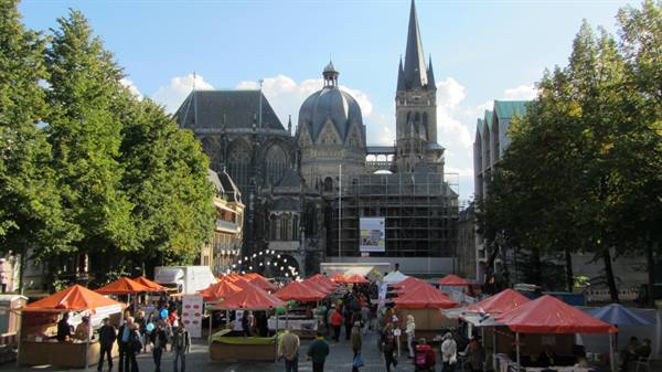 Orange market tents in front of Aachen Cathedral, people strolling among trees