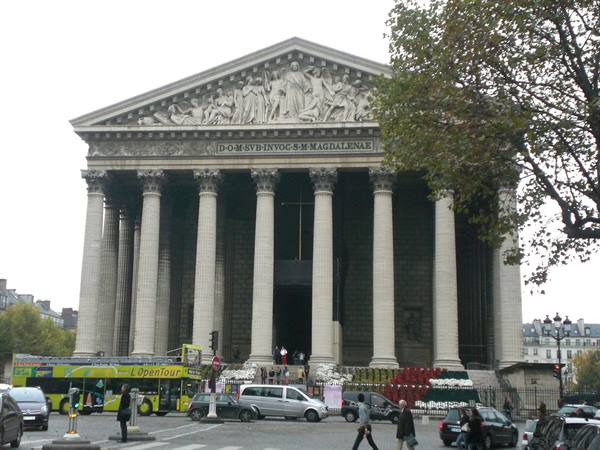 Tourists climb the steps of the Madeleine church among Corinthian columns and traffic