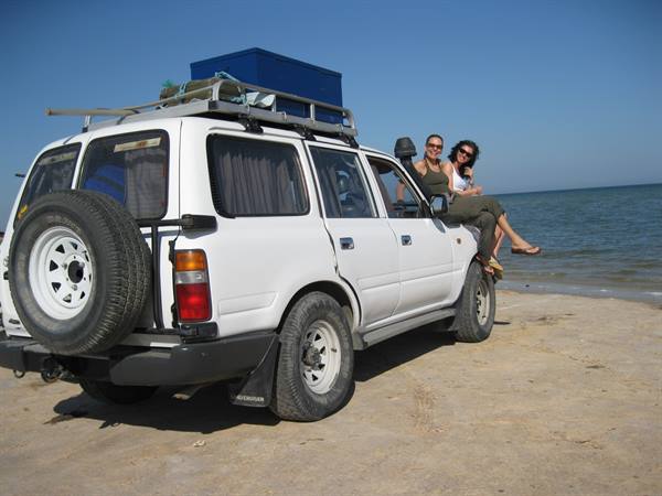 Two women sitting on a white 4x4 parked on a sunny beach
