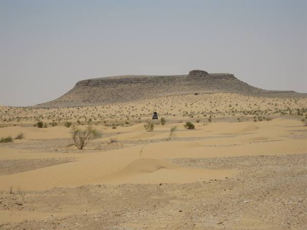Jeep on sandy desert in front of rocky mesa, scattered shrubs